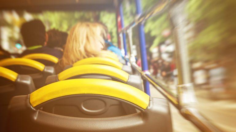 מבט מתוך הסיפון העליון של אוטובוס תיירים בנסיעה: מושבים עם משענות צהובות ונוסעים שמביטים אל הרחוב